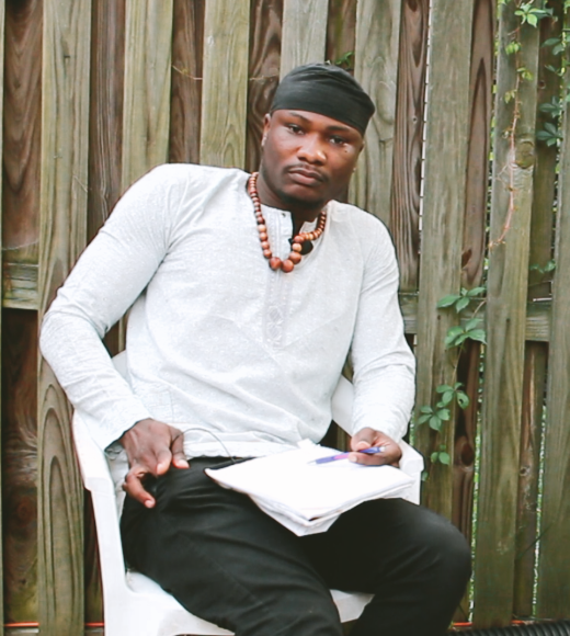 Photo of Ibrahim sitting down, looking straight into the camera. He is wearing white and a beaded necklace. A notebook rests in his lap.