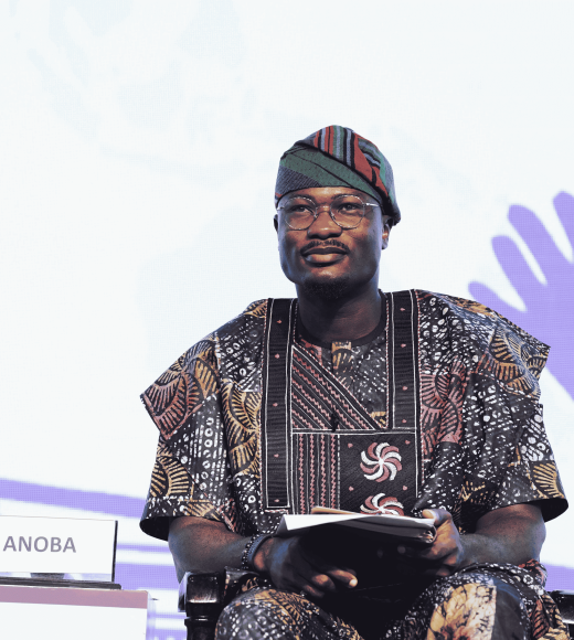 Ibrahim sitting down in chair with a book on his lap and carrying a microphone in one hand. He is wearing glasses and multi-colored cultural garments.