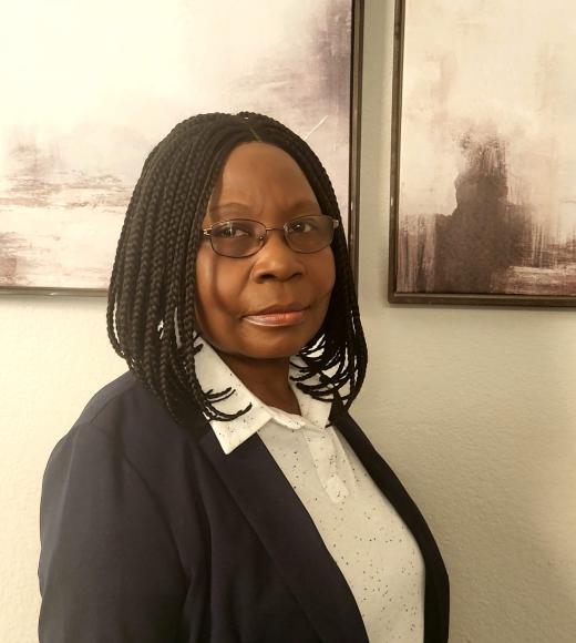 Photo of Elizabeth in a black blazer and a white-collared button down shirt. She is wearing glasses and standing in front of a neutral shade background.