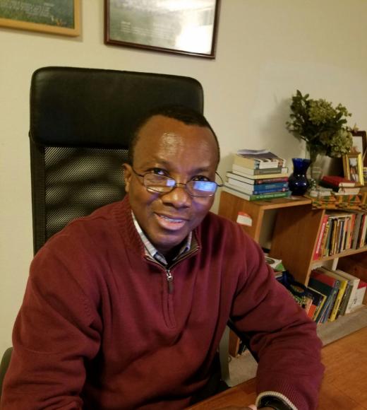 Photo of Tometi sitting down at a table. He has his hands resting on the table in front of him, and he is wearing glasses and a burgundy sweater.