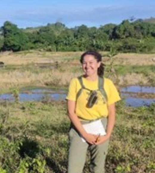 Photo of Marie standing in a field out in a natural environment. She is wearing a yellow T-shirt and light olive green pants. She has a backpack on and is holding a notebook. 