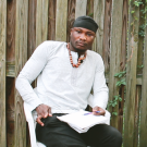 Photo of Ibrahim sitting down, looking straight into the camera. He is wearing white and a beaded necklace. A notebook rests in his lap.