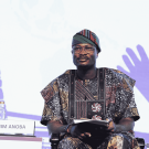 Ibrahim sitting down in chair with a book on his lap and carrying a microphone in one hand. He is wearing glasses and multi-colored cultural garments.