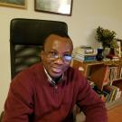 Photo of Tometi sitting down at a table. He has his hands resting on the table in front of him, and he is wearing glasses and a burgundy sweater.