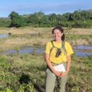 Photo of Marie standing in a field out in a natural environment. She is wearing a yellow T-shirt and light olive green pants. She has a backpack on and is holding a notebook. 